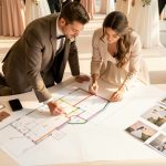 Un jeune couple de futurs mariés prépare un parcours de visite sur un plan vierge, avec un carnet, un téléphone et des brochures élégantes posés sur une table.